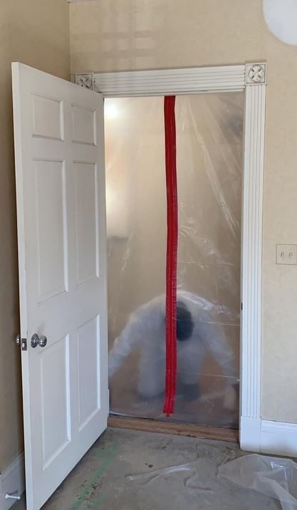 Doorway sealed with plastic sheeting and a zipper door, with a worker in a protective suit inside the containment